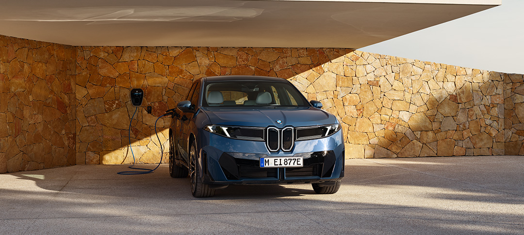 : Ein blauer BMW Elektro-SUV der „Neue Klasse“ Konzeptreihe parkt unter einem modernen Carport und ist über ein Ladekabel an einer Wallbox an einer Natursteinwand angeschlossen.