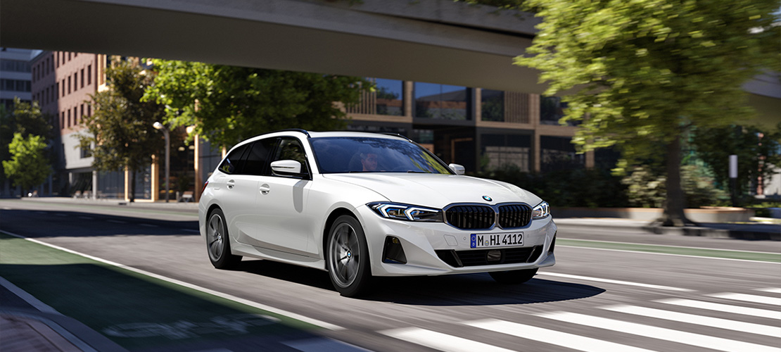 Ein dynamisches Pressefoto zeigt einen weißen BMW 3er Touring während einer zügigen Fahrt durch ein modernes Stadtviertel. Das Fahrzeug wird von vorne links abgebildet und ist mit dem Münchner Kennzeichen M-HI 4112 ausgestattet. Durch leichte Bewegungsunschärfe an den Rädern und der Umgebung wird eine fließende Bewegung erzeugt. Der Hintergrund besteht aus modernen Bürogebäuden, einer erhöhten Betonbrücke und grünen Bäumen unter klarem Tageslicht.