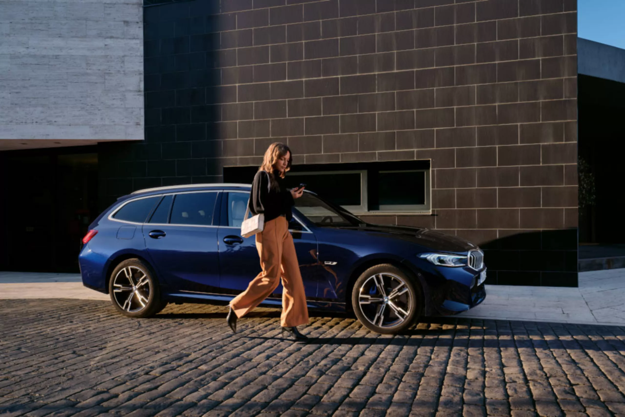 BMW PREMIUM SELECTION GARANTIE Müller Frau geht neben einem dunkelblauen BMW Kombi auf einem gepflasterten Weg.