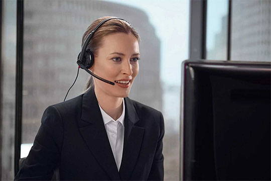 Frau mit Headset und Mikrofon lächelt während eines Gesprächs am Computer in einem Büro.