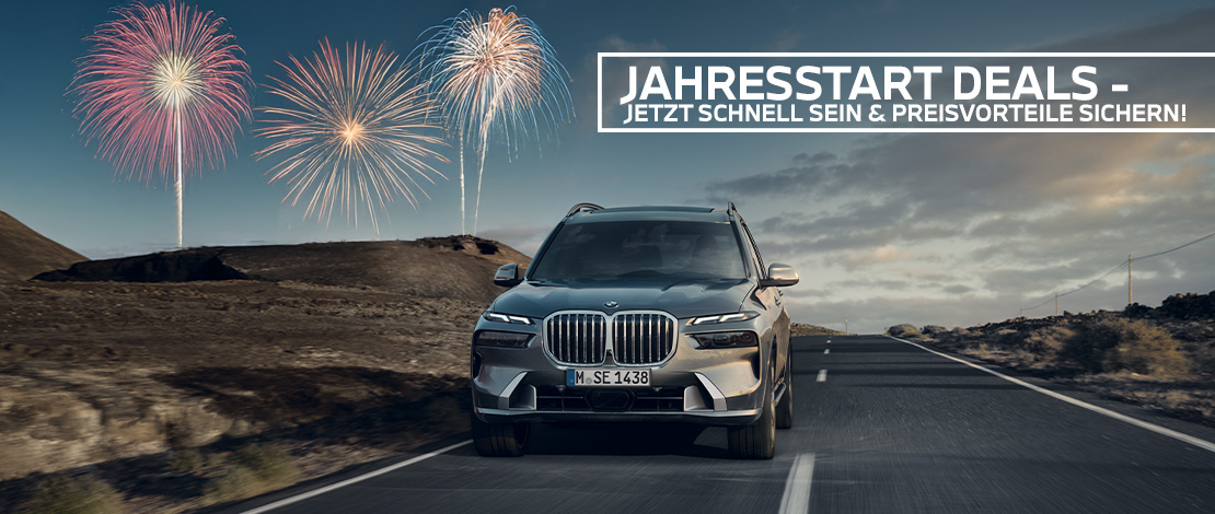 Das Bild ist ein großformatiges Header-Banner für eine Werbeaktion. Es zeigt einen luxuriösen, dunkelgrauen BMW SUV (BMW X7), der frontal auf einer asphaltierten Straße durch eine karge, vulkanähnliche Landschaft fährt. Der Himmel ist in der Dämmerung gehalten und wird von drei großen, farbenfrohen Feuerwerken in den Farben Rosa, Gold und Blau erhellt. Oben rechts befindet sich eine weiße Textbox mit der Aufschrift: „JAHRESSTART DEALS – JETZT SCHNELL SEIN & PREISVORTEILE SICHERN!“. Das Bild vermittelt eine festliche Aufbruchstimmung zum neuen Jahr.