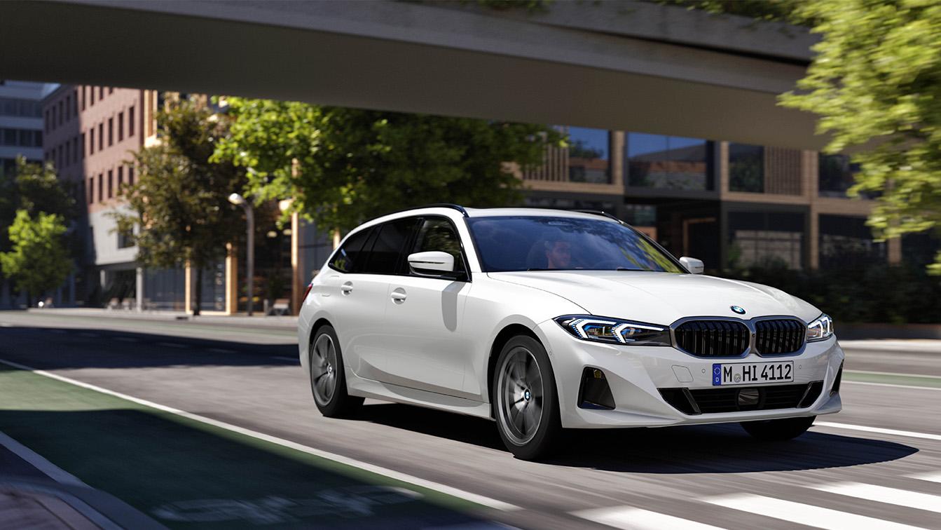 Das Bild zeigt einen weißen BMW 3er Touring, der dynamisch durch eine moderne Stadtkulisse fährt. Die Kombination aus der glänzenden Lackierung, den scharfen LED-Scheinwerfern und der leichten Bewegungsunschärfe auf dem Asphalt vermittelt einen sportlichen und hochwertigen Eindruck. Die Architektur im Hintergrund und das natürliche Sonnenlicht unterstreichen dabei den urbanen, eleganten Charakter des Fahrzeugs.