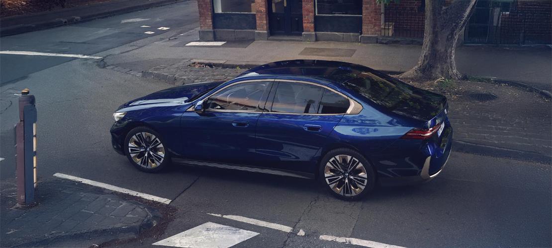 BMW 5er Limousine zum TOP ANGEBOT Eine dunkelblaue BMW 5er Limousine steht auf einer Straße, von oben gesehen. Das Auto ist in einem modernen, eleganten Design gehalten, mit glänzenden Felgen und Chromakzenten. Es ist auf einer leicht abfallenden Straße geparkt, neben einem Gehweg mit einem Baum und einem Gebäude im Hintergrund. Das Licht fällt von oben auf das Auto, wodurch die Linien und Kurven des Fahrzeugs hervorgehoben werden.