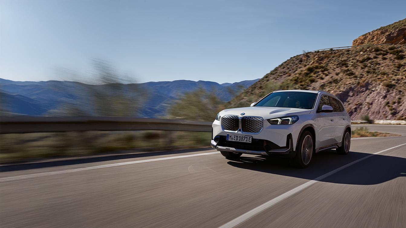 Ein weißer BMW iX1 fährt zügig über eine kurvige Bergstraße unter einem klaren, hellblauen Himmel direkt auf den Betrachter zu. Während das Fahrzeug in der rechten Bildhälfte gestochen scharf abgebildet ist, erzeugen der verwischte Asphalt und die vorbeiziehenden Leitplanken ein starkes Gefühl von Dynamik und Geschwindigkeit. Die markante Front des Elektro-SUVs mit dem geschlossenen, hellen Kühlergrill und den präzisen LED-Scheinwerfern steht im Fokus, umrahmt von einer weitläufigen Landschaft mit bläulichen Bergketten im Hintergrund und einem kargen, sonnenbeschienenen Felshang zur Rechten.