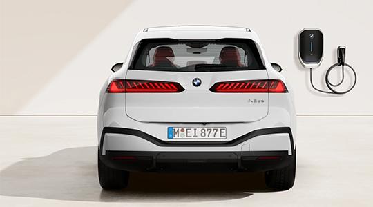 Auf dem Bild ist die Rückansicht eines weißen BMW iX3 zu sehen. Das Auto steht in einem Studio, und rechts davon ist eine schwarze BMW Wallbox, eine Ladestation, an der Wand befestigt. Die breiten, zweigeteilten Heckleuchten des Autos sind leuchtend rot und haben ein auffälliges, gestreiftes Design. In der Mitte der Heckklappe befindet sich das BMW-Emblem. Unterhalb des Nummernschilds ist eine dezente schwarze Kunststoffverkleidung. Durch die Heckscheibe sind die roten Sitze im Innenraum zu erkennen.