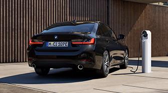 BMW Plug-in-Hybride Das Bild zeigt die Heckansicht eines schwarzen BMW 3er Plug-in-Hybrid. Das Auto steht draußen vor einer modernen Hausfassade mit Holzverkleidung. Die Rückleuchten sind an. Rechts neben dem Auto befindet sich eine Ladesäule mit einem angeschlossenen Kabel, das in das Auto führt.