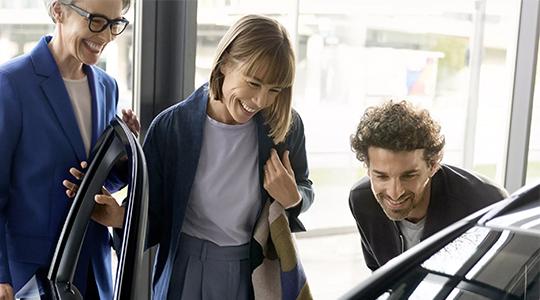 BMW Plug-in-Hybrid Neuwagen Das Bild zeigt drei Personen in einem Autohaus, die sichtlich fröhlich sind. Sie stehen um ein Auto herum, das nur teilweise zu sehen ist. Eine Frau in einem blauen Blazer, die eine Brille trägt, lacht und schaut in den Innenraum des Autos. Eine andere Frau mit kurzen Haaren in einer blauen Jacke schaut ebenfalls lächelnd in den Innenraum des Wagens. Ein Mann mit lockigen Haaren beugt sich ebenfalls lachend über die Motorhaube des Autos. Die Atmosphäre wirkt sehr positiv und entspannt.