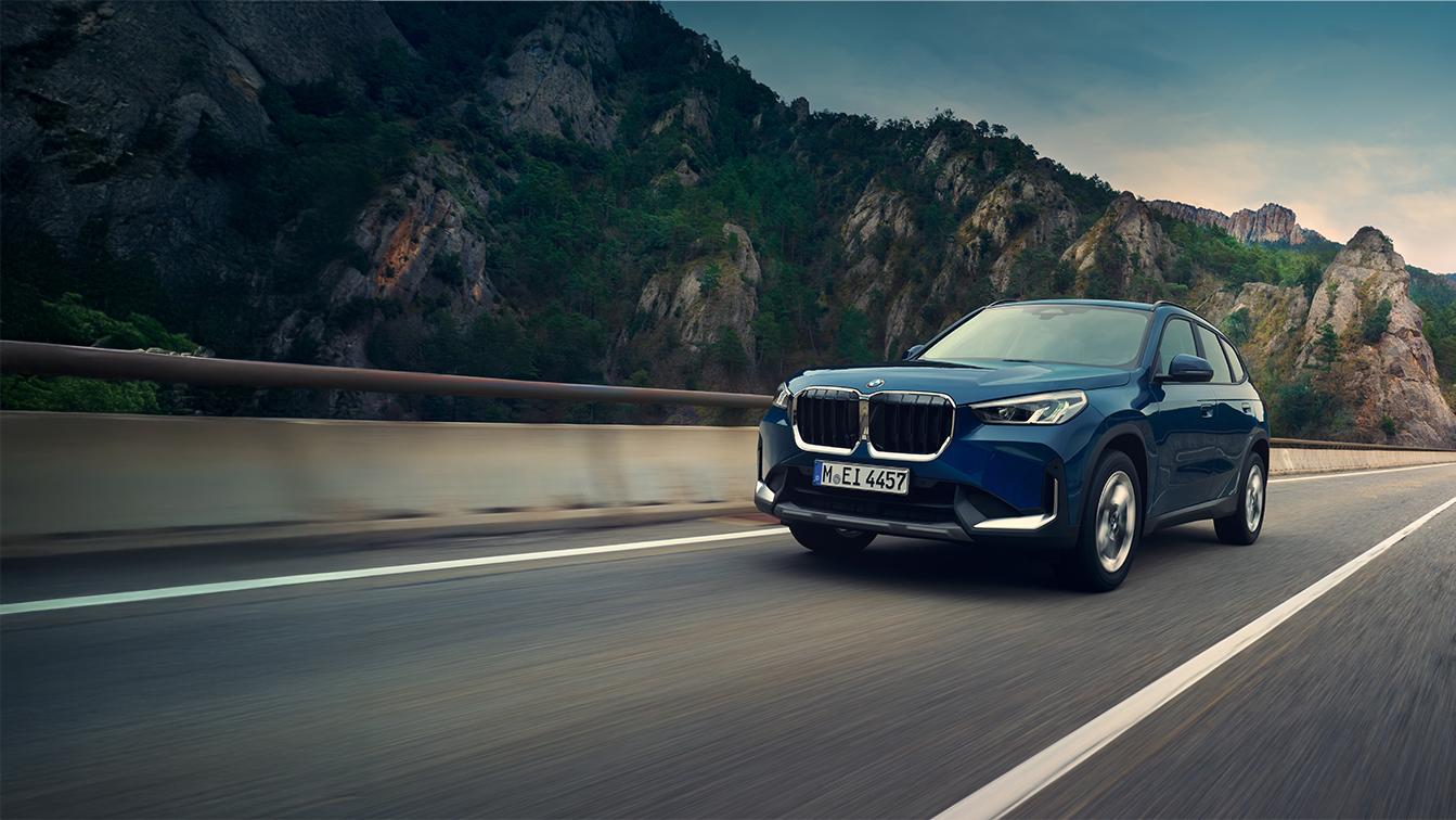Ein dunkelblauer BMW X1 SUV fährt dynamisch auf einer asphaltierten Straße entlang einer Leitplanke vor einer imposanten, steilen Bergkulisse.