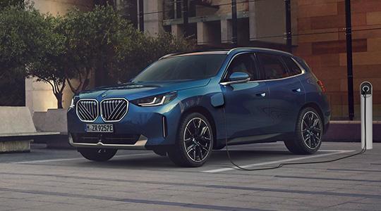 Das Bild zeigt den dunkelblauen BMW X3 von vorne links auf einem gepflasterten Parkplatz vor einem modernen Gebäude. Das Fahrzeug ist über ein schwarzes Kabel an einer minimalistischen, weißen Ladesäule angeschlossen, wobei die geöffnete Ladeklappe am vorderen linken Kotflügel gut sichtbar ist.