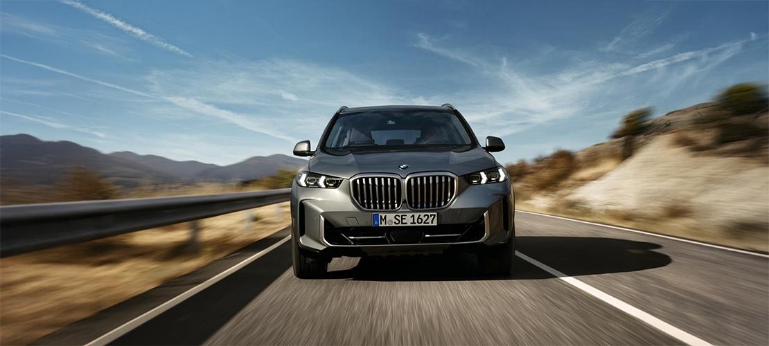 BMW X5 Angebot Ein dunkelgrauer oder silberner BMW X5 SUV fährt auf einer kurvigen Landstraße auf den Betrachter zu. Das Auto hat die charakteristische BMW-Niere und eingeschaltete Scheinwerfer. Im Hintergrund sind Berge und ein klarer blauer Himmel zu sehen, was auf eine Fahrt durch eine malerische Landschaft hindeutet. Das Kennzeichen lautet "M SE 1627".