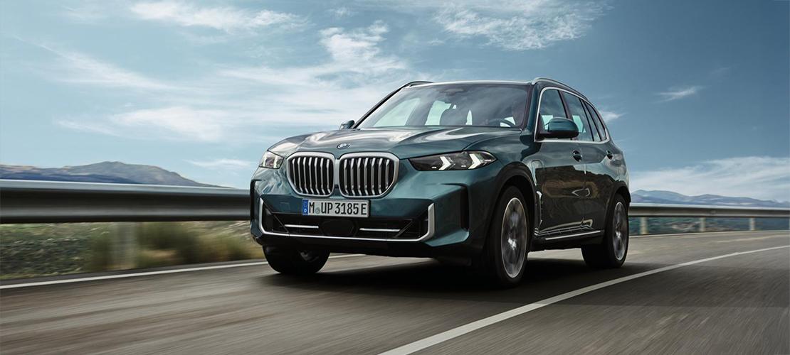  Ein Plug-in Hybrid BMW X5 in einer dunklen Petrol- oder Grünmetallic-Farbe fährt auf einer mehrspurigen Straße. Das Fahrzeug ist dynamisch von vorne-rechts aufgenommen, wodurch die markante BMW-Niere, die schmalen Scheinwerfer und die sportlichen Linien gut zur Geltung kommen. Die Landschaft im Hintergrund besteht aus verschwommenen Bergen und einem weiten Himmel mit einigen Wolken, was den Eindruck von Geschwindigkeit und Bewegung verstärkt. Das Kennzeichen des Fahrzeugs ist "M UP 3185 E".
