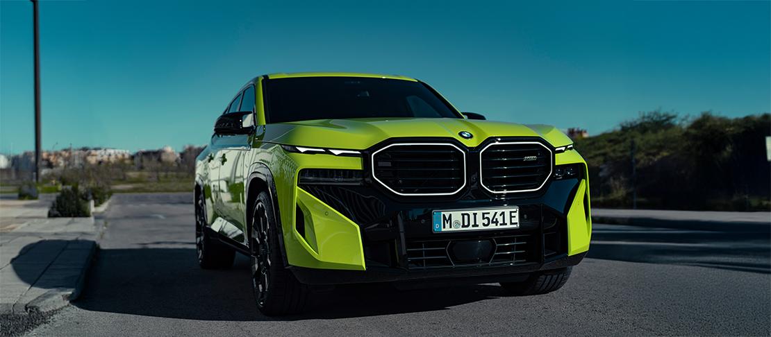 Ein markanter, neongelber BMW XM dominiert die Szene. Die Frontalansicht hebt die beleuchteten, eckigen Niere-Grills und die aggressiven Lufteinlässe hervor. Mit seinem deutschen E-Kennzeichen parkt der Performance-SUV auf einer Straße vor einer modernen, industriellen Kulisse unter strahlend blauem Himmel.