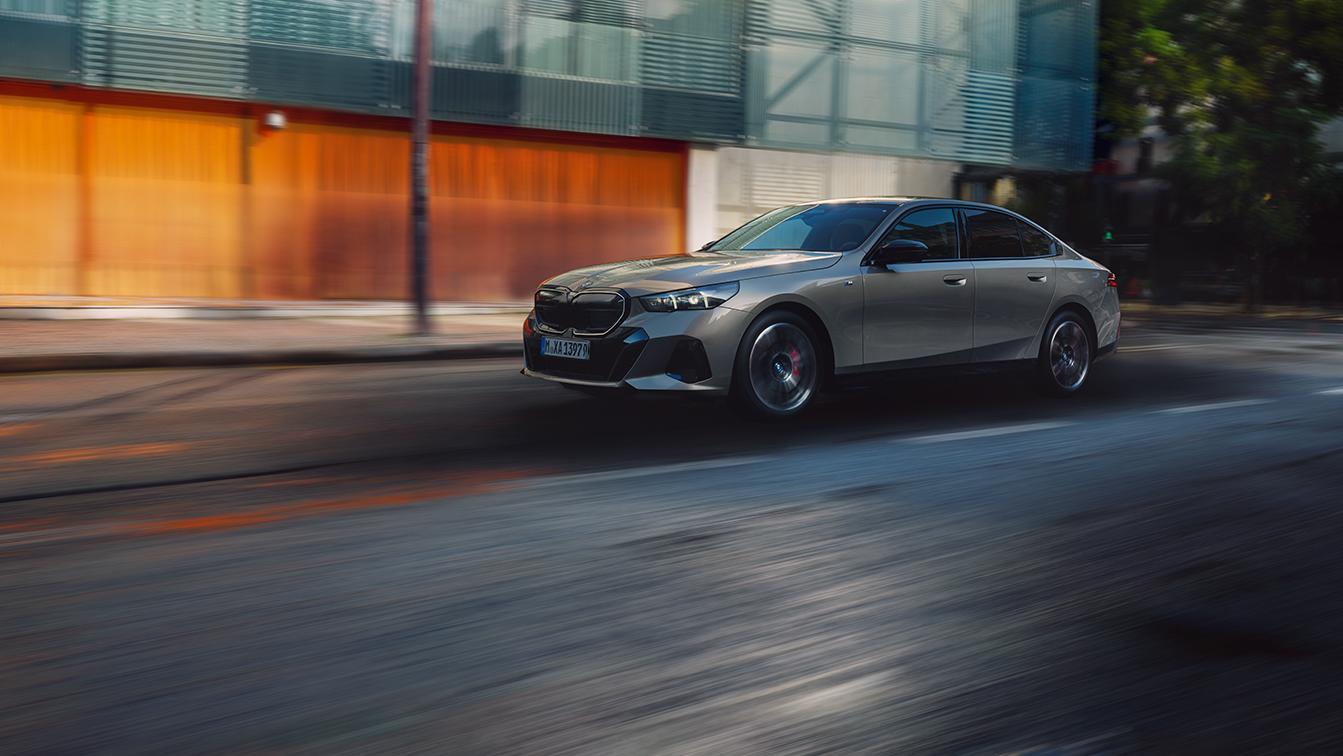  Ein silberner BMW 5er (G60) fährt dynamisch durch eine urbane Kulisse. Die starke Bewegungsunschärfe betont die Geschwindigkeit, während das Fahrzeug vor einer modernen Glasfassade und warmen, orangefarbenen Akzenten im Hintergrund kontrastreich in Szene gesetzt wird.