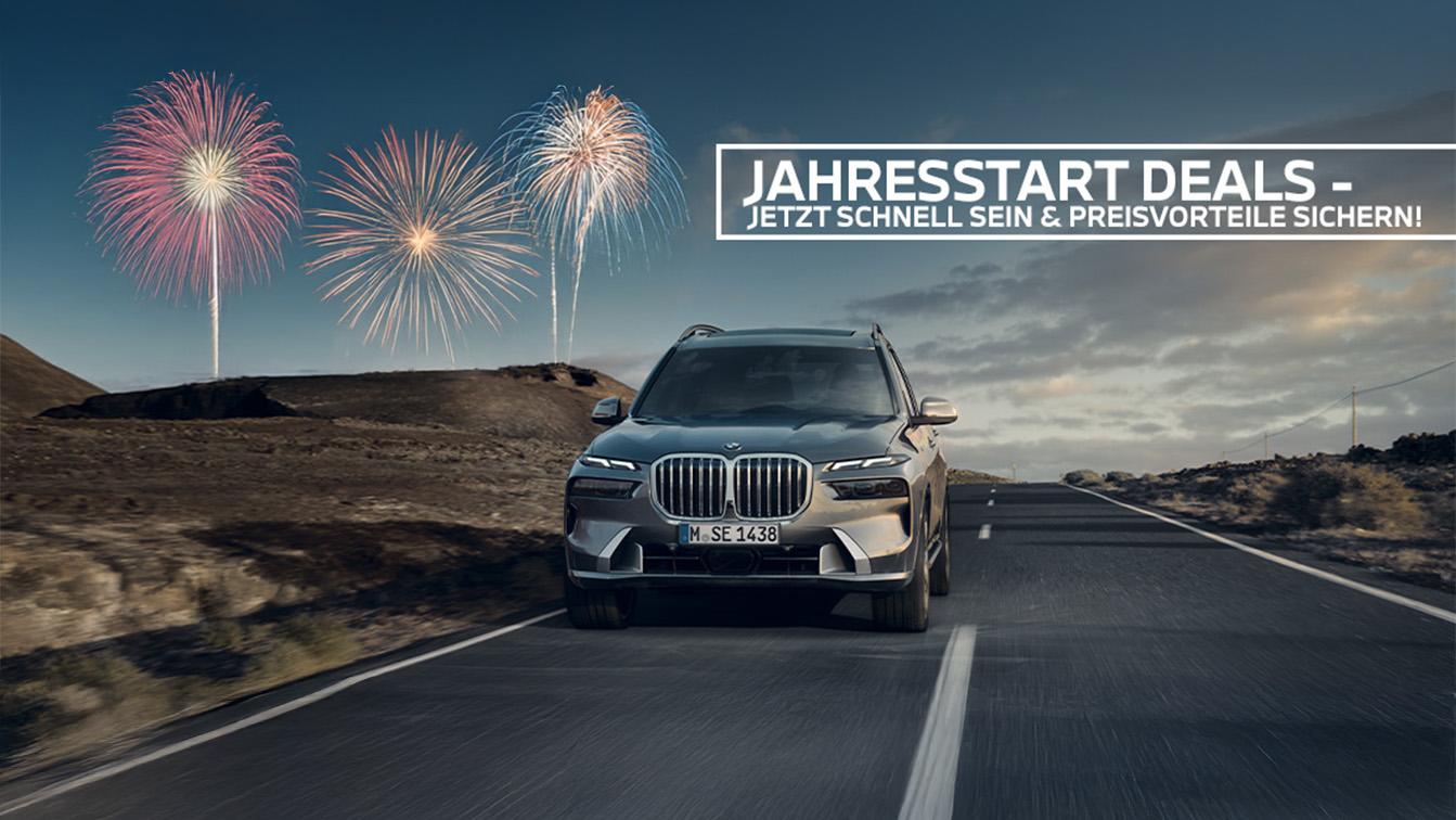 Das Bild ist ein horizontales Werbebanner für ein BMW-Autohaus zum Thema „Jahresstart Deals“. Es zeigt einen luxuriösen, silbernen BMW SUV (Modell X7), der auf einer asphaltierten Landstraße durch eine karge, hügelige Landschaft fährt. Am dämmerigen Himmel über der Szenerie steigen feierliche Feuerwerke auf. Oben rechts befindet sich ein weißer Textkasten mit der Aufschrift: „JAHRESSTART DEALS – JETZT SCHNELL SEIN & PREISVORTEILE SICHERN!“.