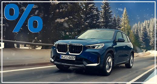 Ein dunkelblauer BMW X1 SUV fährt auf einer verschneiten Straße. Im Hintergrund erstrecken sich dichte, verschneite Tannenwälder. Das Auto ist leicht von vorne rechts aufgenommen. Das blaue Prozentzeichen (%) füllt die obere linke Ecke und signalisieren einen Rabatt oder eine spezielle Finanzierung.
