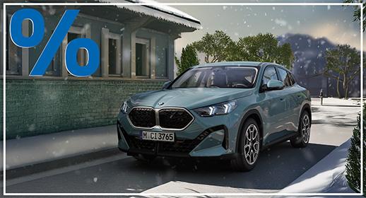 Dieses Bild zeigt einen türkis- oder mintgrünen BMW X2, der auf einer verschneiten Straße oder einem Weg geparkt ist oder langsam fährt. Im Hintergrund links ist das Detail eines Gebäudes mit dunkelgrün-blauer Fassade und einem schneebedeckten Dach zu sehen, möglicherweise ein Bahnhof oder ein traditionelles Haus. Rechts und im Hintergrund befinden sich Bäume und eine verschneite Landschaft mit angedeuteten Bergen. Die Szene ist winterlich, mit leichtem Schneefall. Ähnlich wie beim ersten Bild, dominiert in der oberen linken Ecke ein großes, blaues Prozentzeichen (%), das auf ein aktuelles Verkaufs- oder Finanzierungsangebot hinweist.