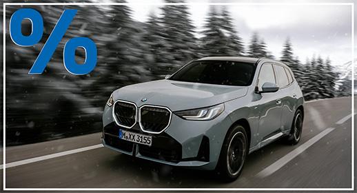 Ein hellgrauer BMW X3 SUV ist von vorne links auf einer verschneiten Straße zu sehen, die an einem Wald mit verschneiten Bäumen vorbeiführt. Das Fahrzeug bewegt sich schnell, was durch die Bewegungsunschärfe im Hintergrund angedeutet wird. Das blaue Prozentzeichen (%) füllt die obere linke Ecke und signalisieren einen Rabatt oder eine spezielle Finanzierung.