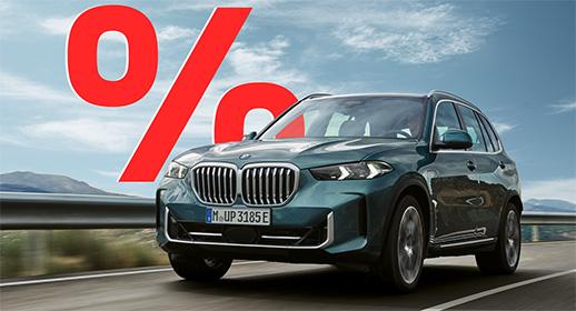 Das Bild zeigt einen dunkelgrünen BMW X5, der auf einer Landstraße fährt. Das Auto ist von vorne zu sehen. Im Vordergrund, über dem Auto, ist ein großes, rotes Prozentzeichen (%) platziert. Der Hintergrund zeigt eine bergige Landschaft unter einem blauen Himmel mit einigen Wolken. Das Kennzeichen "M UP 3185 E" ist auf der vorderen Stoßstange zu sehen.