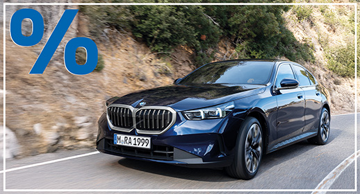 Das Bild zeigt eine dunkelblaue, viertürige BMW 5er-Reihe Limousine, die auf einer kurvigen Straße fährt. Das Auto ist von vorne zu sehen. Im Vordergrund ist ein großes, rotes Prozentzeichen (%) platziert. Der Hintergrund zeigt eine bergige Landschaft mit Bäumen und Sträuchern. Das Kennzeichen "M RA 1999" ist auf der vorderen Stoßstange zu sehen.
