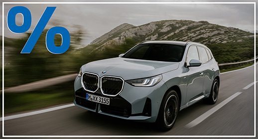 Das Bild zeigt einen hellgrauen, viertürigen BMW X3, der auf einer Landstraße fährt. Das Auto ist schräg von vorne zu sehen. Im Vordergrund ist ein großes, rotes Prozentzeichen (%) platziert. Der Hintergrund zeigt eine bergige Landschaft unter einem bewölkten Himmel. Das Kennzeichen "M XX 3155" ist auf der vorderen Stoßstange zu sehen.