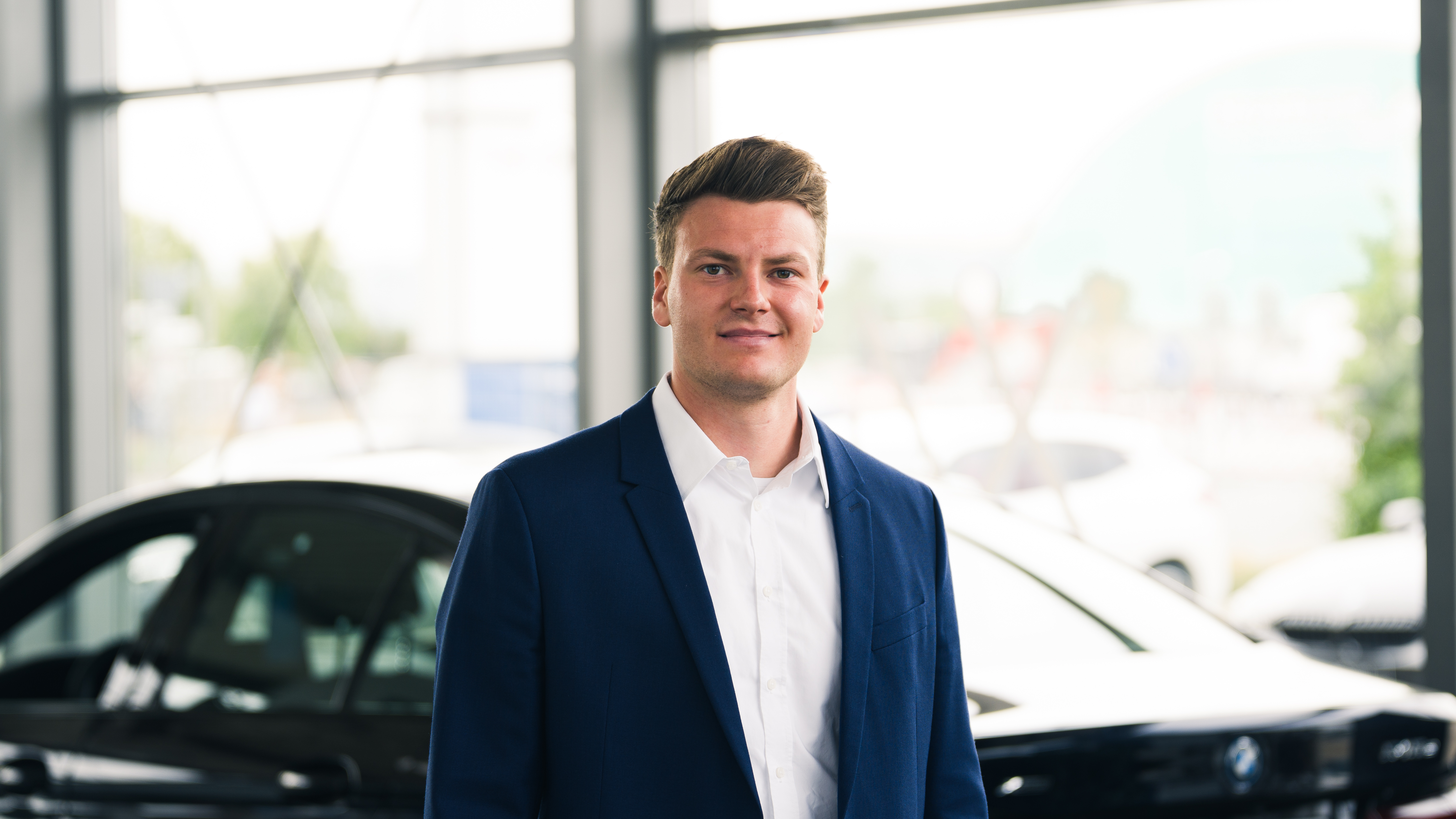 Ein junger Mann mit kurzen braunen Haaren, gekleidet in einen dunkelblauen Anzug und ein weißes Hemd, steht in einem Autohaus und blickt lächelnd in die Kamera. Im unscharfen Hintergrund sind die Umrisse von Autos und große Fenster zu sehen